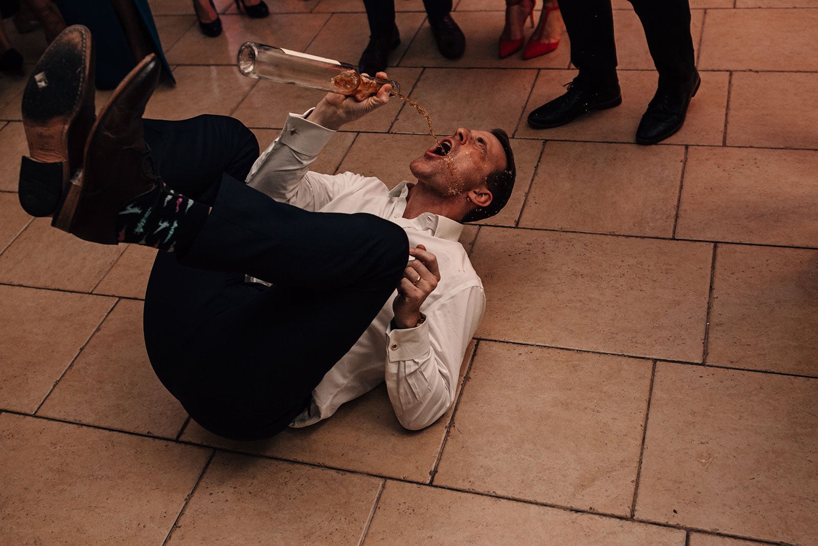 Funny photograph of a man laid down pouring alcohol onto his face on the wedding dance-floor