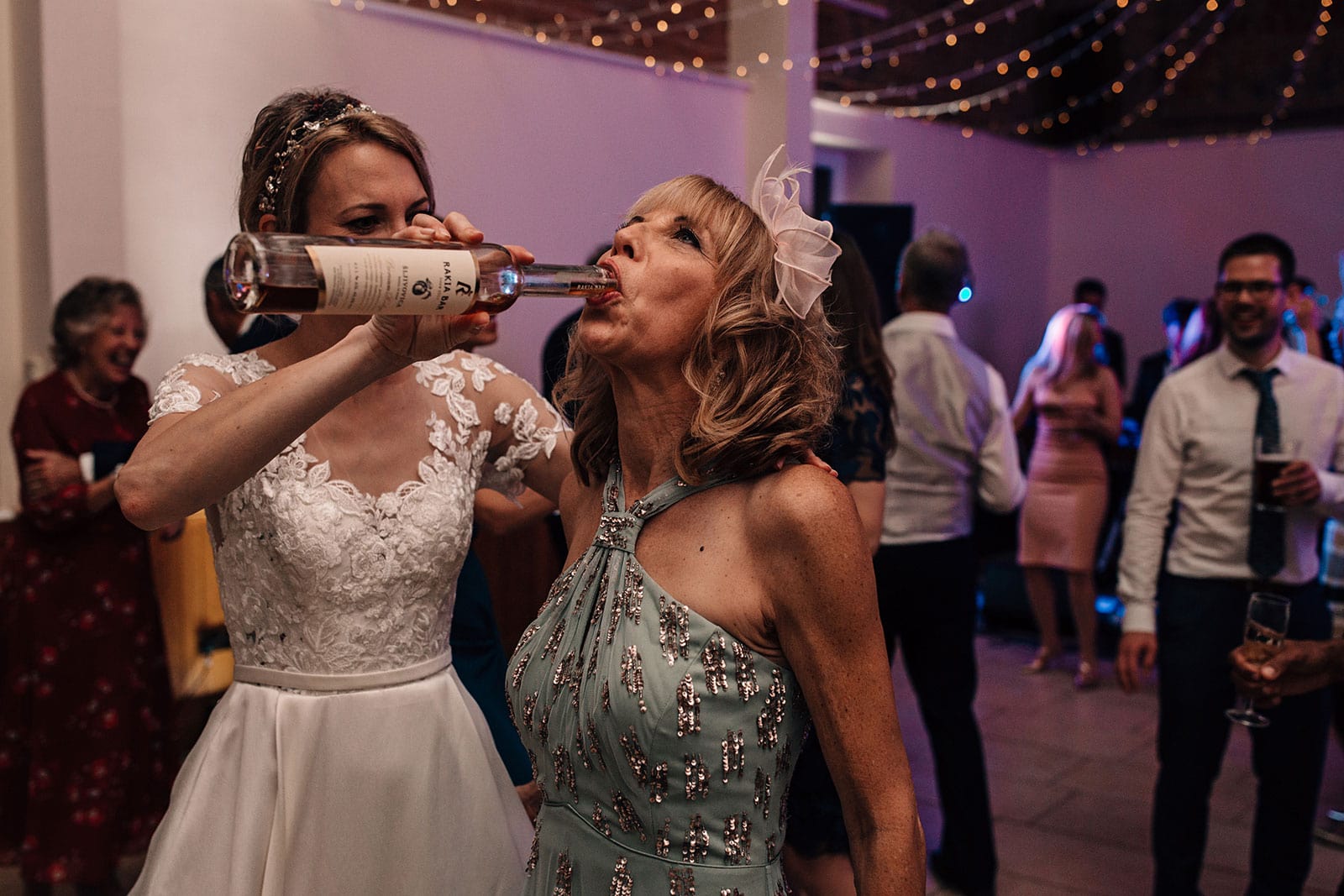 Lively and fun wedding photography of the Bride pouring a bottle of drink into her mothers mouth on the wedding dance-floor