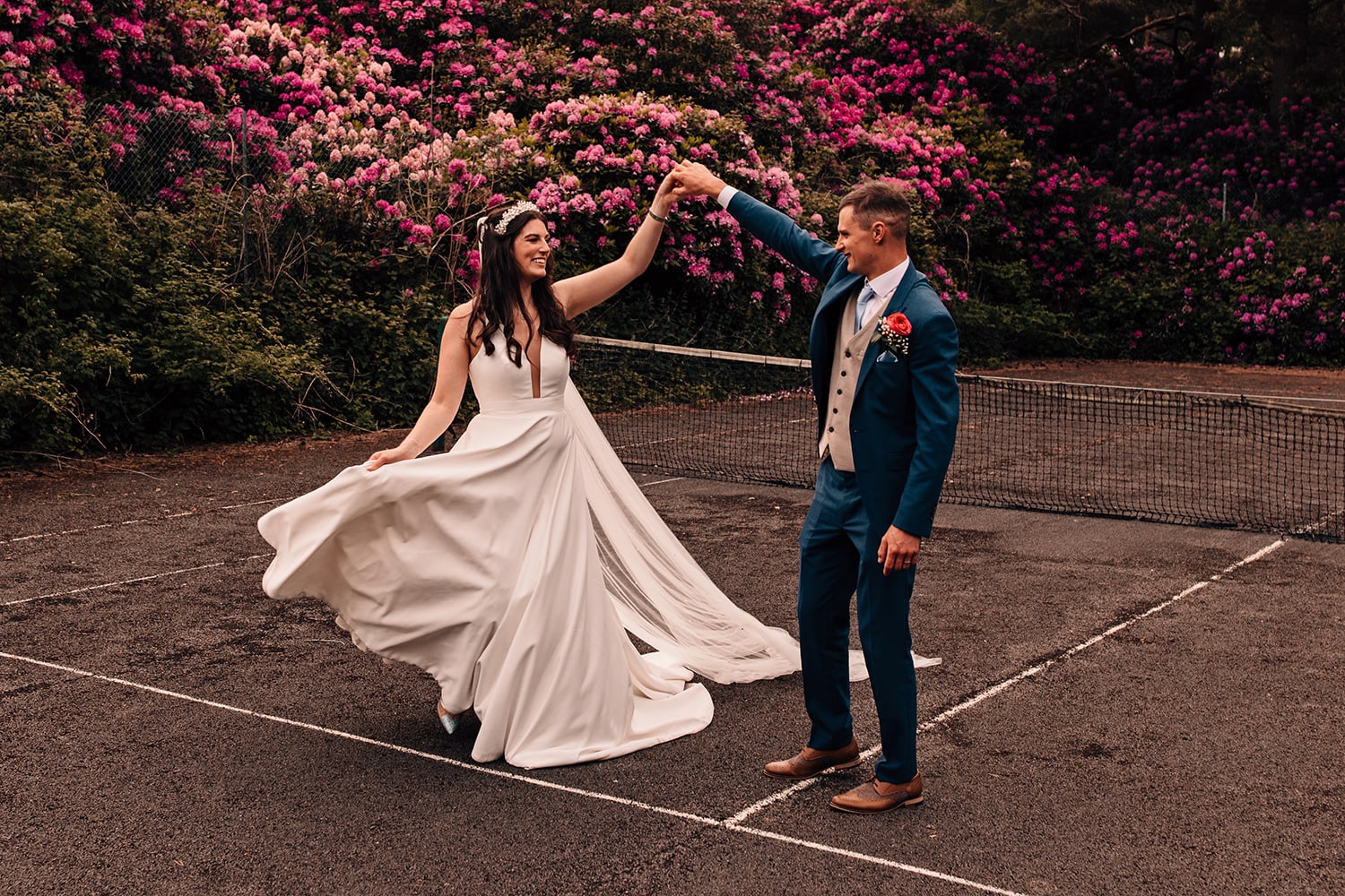 Bride and groom dancing on an abandoned tennis court infront of a large rhododendron bush