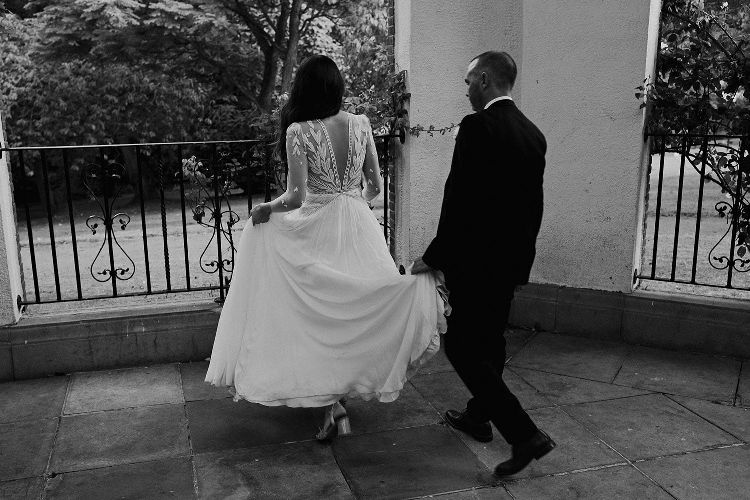Artistic black and white image of a bride walking and her Groom carrying the train of her dress