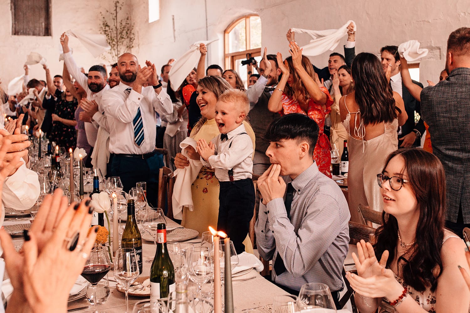 All the wedding guests waving their napkins, cheering and standing on their chairs welcoming the couple into the room