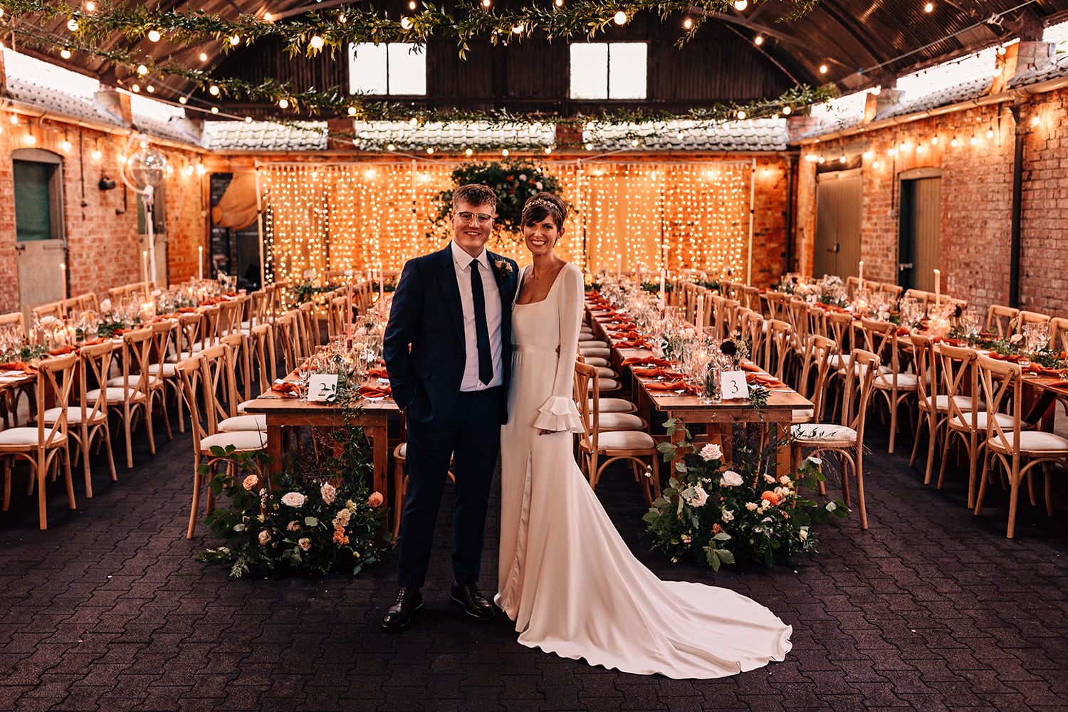 Bride and Groom stood in front of a wall of fairy lights and their rustic barn wedding table set up